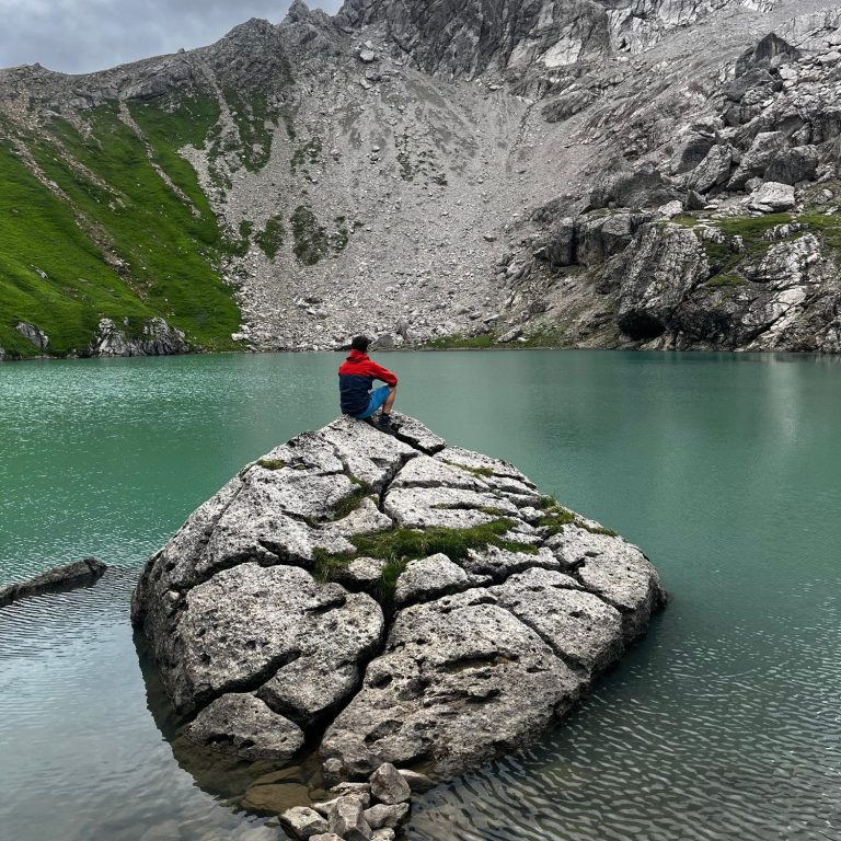 Beratung Person in rotem Pullover sitzt auf einem großen Stein am Ufer eines klaren Sees, umgeben von Bergen.
