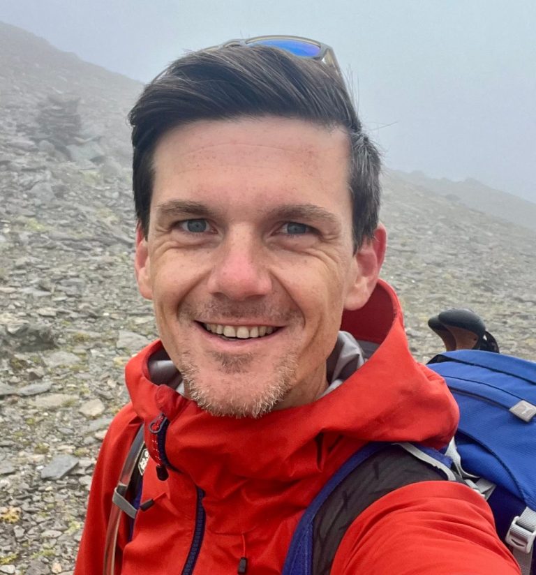 Sebastian Wiedemann Gründer von explore and elevate Mann mit roter Jacke und Rucksack, lächelt in einer bergigen, nebligen Landschaft.