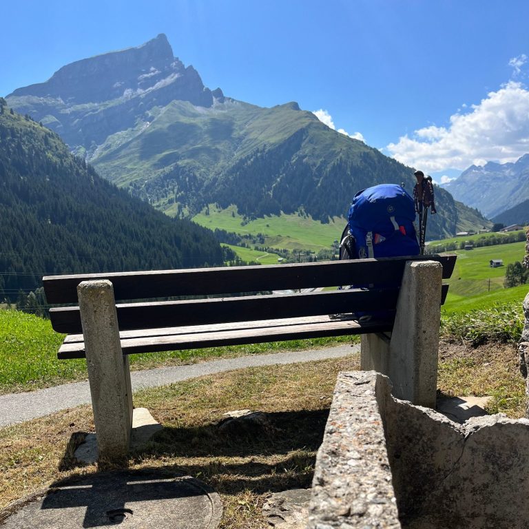 Bank mit Rucksack Blick auf eine Bank in den Alpen mit grünen Wiesen und Bergen im Hintergrund.